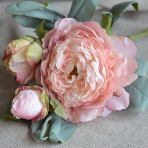 May include: A bouquet of three pink artificial peonies with green leaves. The peonies are in various stages of bloom, with one fully open, one partially open, and one bud.