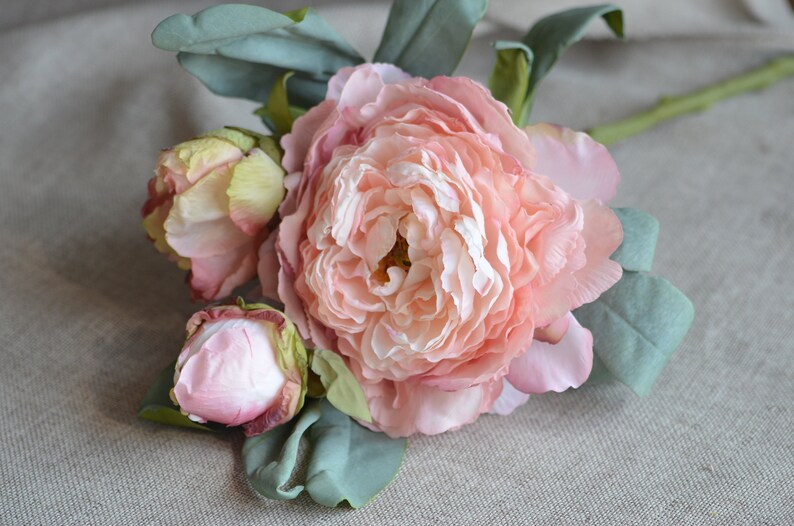 May include: A bouquet of three pink artificial peonies with green leaves. The peonies are in various stages of bloom, with one fully open, one partially open, and one bud.