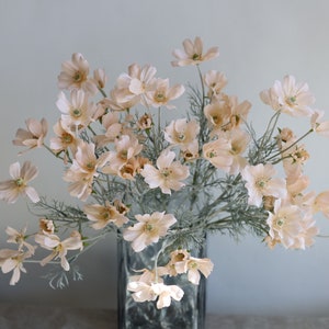 Könnte beinhalten: Ein Strauß künstlicher, blassrosa Blüten mit grünen Stielen in einer durchsichtigen Glasvase. Die Blumen haben einen zarten, femininen Look.