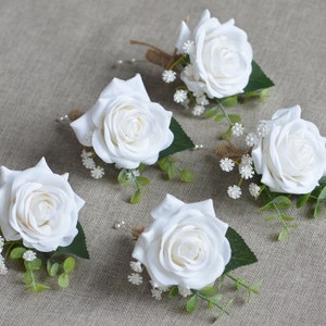 Op de afbeelding: Vijf witte rozen corsages met groen en kleine witte bloemen. Elke corsage heeft een jute strik en een speld.