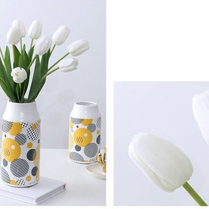 May include: White tulip bouquet in a white ceramic vase with a yellow and gray geometric pattern. The vase is on a white surface with a book.