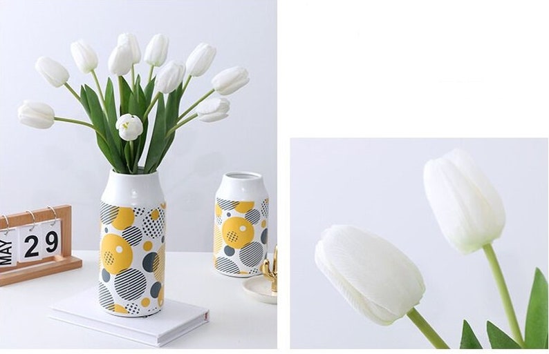 May include: White tulip bouquet in a white ceramic vase with a yellow and gray geometric pattern. The vase is on a white surface with a book.