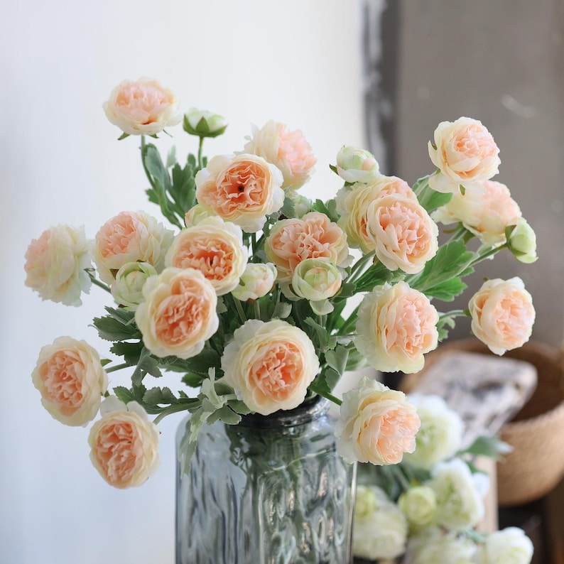 Puede incluir: Un ramo de flores artificiales de color melocot&oacute;n en un jarr&oacute;n de vidrio transparente. Las flores tienen un aspecto delicado y con volantes y est&aacute;n dispuestas en un estilo suelto y natural.