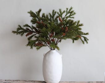 May include: A white ceramic vase with a textured surface holds a lush arrangement of evergreen branches and pine cones. The vase has a rounded shape with two small handles on the sides. The arrangement is a natural, rustic home decor item.