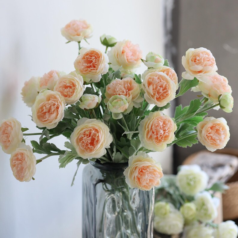 Puede incluir: Un ramo de rosas artificiales de color melocot&oacute;n en un jarr&oacute;n de vidrio transparente. Las rosas est&aacute;n dispuestas en un estilo suelto y natural, con algunos tallos que se extienden m&aacute;s all&aacute; del jarr&oacute;n. El jarr&oacute;n est&aacute; sobre una mesa con un fondo blanco.