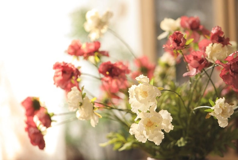 May include: A bouquet of artificial flowers featuring a mix of deep red and creamy white blossoms. The flowers have layered, ruffled petals and are attached to thin green stems with small green leaves, creating a dense, multi-layered floral arrangement.