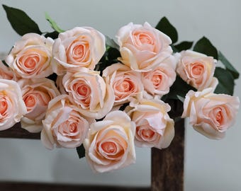 May include: A bouquet of peach-colored artificial roses with green leaves. The roses are arranged in a cluster and are sitting on a wooden surface.
