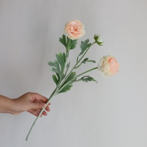 Puede incluir: Un ramo de tres flores de ran&uacute;nculo artificiales de color melocot&oacute;n con hojas verdes.