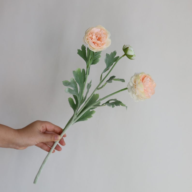 Puede incluir: Un ramo de tres flores de ran&uacute;nculo artificiales de color melocot&oacute;n con hojas verdes.