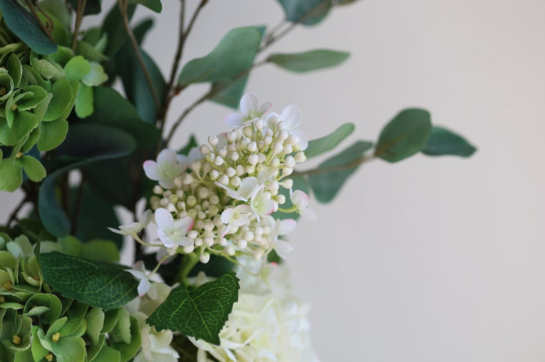 Op de afbeelding: Een boeket witte en groene bloemen met kleine witte bloesems en groene bladeren.