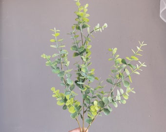 Può includere: Un mazzo di rametti di eucalipto artificiali con foglie verdi. I gambi sono marroni e le foglie sono di colore verde chiaro.