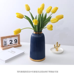 May include: A blue ceramic vase with gold trim and a ribbed design, filled with yellow tulips. The vase is sitting on a white surface with a wooden calendar and a small gold cactus on a gold tray.