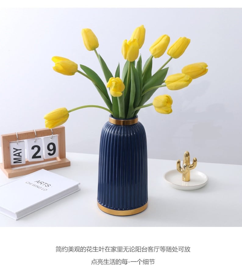 May include: A blue ceramic vase with gold trim and a ribbed design, filled with yellow tulips. The vase is sitting on a white surface with a wooden calendar and a small gold cactus on a gold tray.