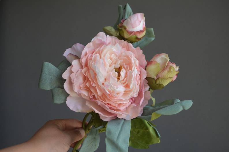 May include: A bouquet of artificial pink peonies with green leaves. The flowers are in various stages of bloom, with one fully open and two buds.