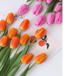 May include: Two bunches of artificial tulips, one bunch is orange and the other is pink. The tulips are arranged on a white surface with a card with a black and white design.