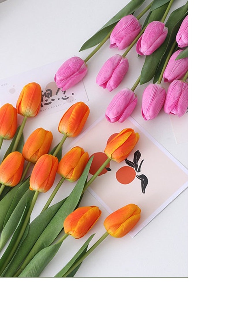May include: Two bunches of artificial tulips, one bunch is orange and the other is pink. The tulips are arranged on a white surface with a card with a black and white design.