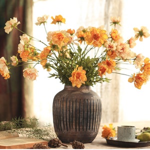 May include: A dark brown, vertically ribbed ceramic vase filled with a large bouquet of artificial flowers in shades of orange, yellow, and cream. The flowers feature ruffled petals and green stems, creating a full, rounded arrangement.