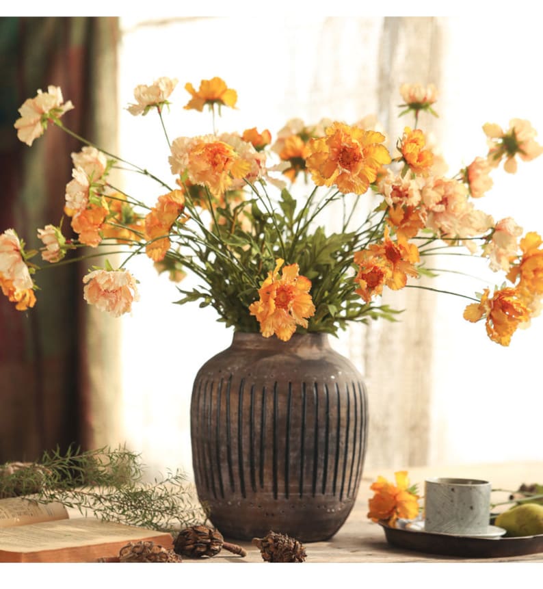 May include: A dark brown, vertically ribbed ceramic vase filled with a large bouquet of artificial flowers in shades of orange, yellow, and cream. The flowers feature ruffled petals and green stems, creating a full, rounded arrangement.