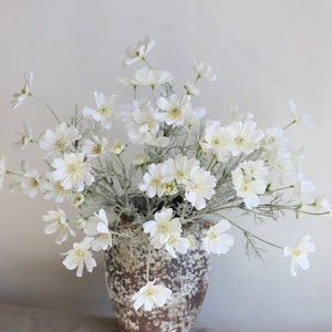 以下が含まれることがあります： 茶色の陶器の花瓶に白い造花の花束。花は繊細な花びらで、緩やかな自然なスタイルにアレンジされています。