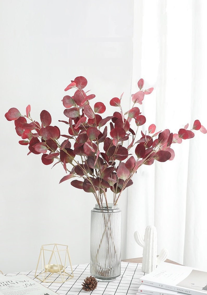 May include: A bouquet of artificial red eucalyptus leaves in a clear glass vase with a ribbed texture. The vase is on a white and black checkered table with a small gold metal stand and a pine cone.