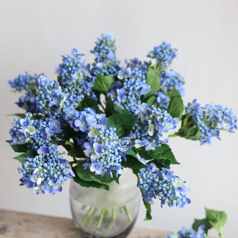 May include: A bouquet of blue artificial hydrangea flowers in a clear glass vase. The flowers are arranged in a cluster and have a realistic appearance.