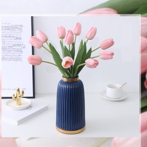 May include: A bouquet of pink tulips in a blue and gold striped vase. The vase is on a white surface with a cactus and a music book in the background.