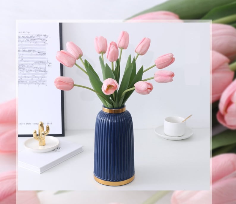 May include: A bouquet of pink tulips in a blue and gold striped vase. The vase is on a white surface with a cactus and a music book in the background.