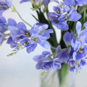 Puede incluir: Un ramo de flores de freesia moradas con centros amarillos en un jarr&oacute;n de vidrio transparente.