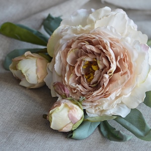 May include: A bouquet of three blush-coloured peonies with green leaves. The peonies are arranged on a light brown linen fabric.
