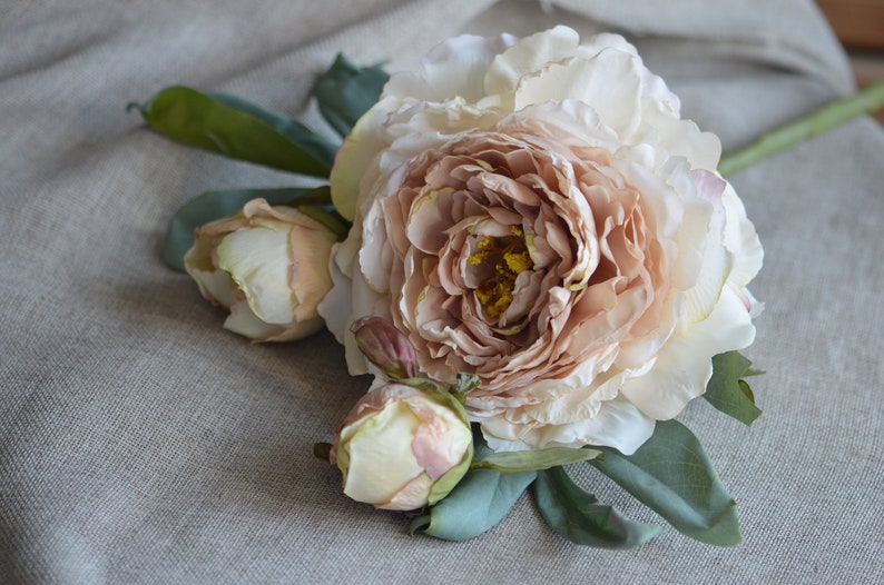 May include: A bouquet of three blush-coloured peonies with green leaves. The peonies are arranged on a light brown linen fabric.