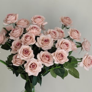 Puede incluir: Un ramo de rosas artificiales de color rosa claro en un jarr&oacute;n de vidrio transparente. Las rosas est&aacute;n dispuestas en un estilo suelto y natural, con algunos tallos que se extienden m&aacute;s all&aacute; del jarr&oacute;n.