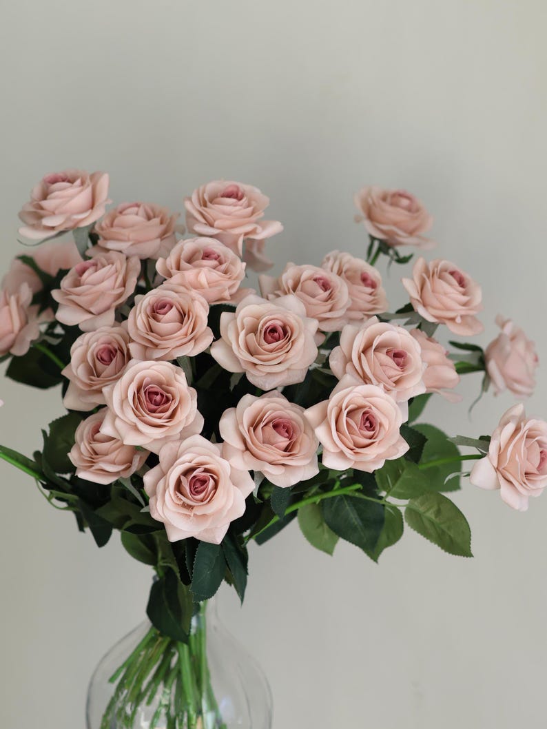 Puede incluir: Un ramo de rosas artificiales de color rosa claro en un jarr&oacute;n de vidrio transparente. Las rosas est&aacute;n dispuestas en un estilo suelto y natural, con algunos tallos que se extienden m&aacute;s all&aacute; del jarr&oacute;n.