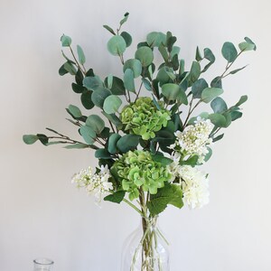 Op de afbeelding: Een heldere glazen vaas met een getextureerd oppervlak bevat een boeket groene en witte bloemen. De bloemen omvatten eucalyptusbladeren en witte hortensia's.