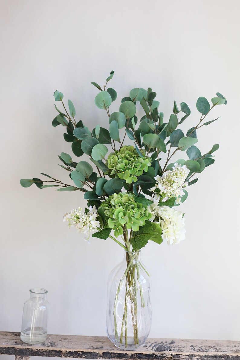 Op de afbeelding: Een heldere glazen vaas met een getextureerd oppervlak bevat een boeket groene en witte bloemen. De bloemen omvatten eucalyptusbladeren en witte hortensia's.