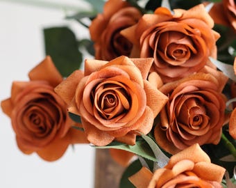 May include: A bouquet of artificial roses in a deep orange color. The roses are arranged in a cluster, with the petals overlapping and creating a soft, romantic look.