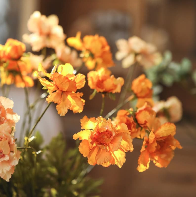 May include: A cluster of artificial flowers featuring vibrant orange and soft peach-colored petals with ruffled edges. The blooms are attached to thin green stems with small green leaves, creating a dense arrangement of multiple flower heads.