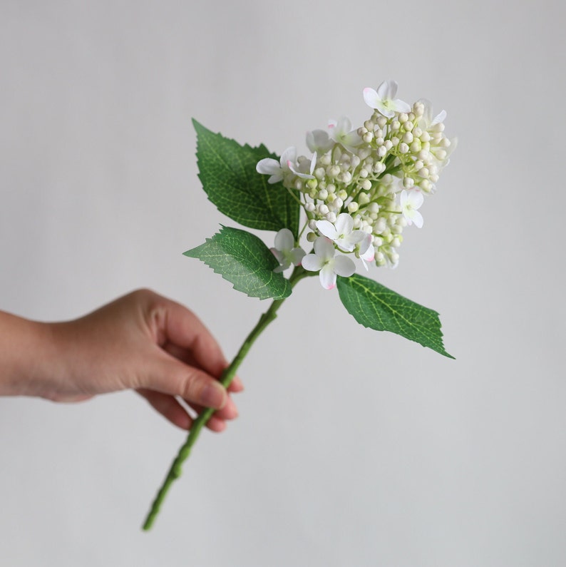 May include: A single stem of white artificial hydrangea with green leaves. The flowers are clustered together and have a delicate, realistic appearance.