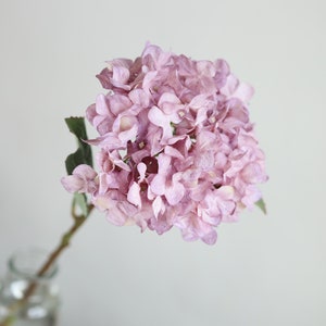 Puede incluir: Un solo tallo de flores de hortensia artificiales de color lila claro. Las flores est&aacute;n agrupadas en forma redonda.