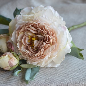 May include: A bouquet of artificial peonies with a large, open bloom in a peachy pink colour and two smaller buds in a cream colour. The flowers are arranged on a green stem with leaves.