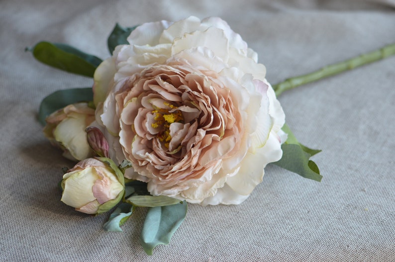 May include: A bouquet of artificial peonies with a large, open bloom in a peachy pink colour and two smaller buds in a cream colour. The flowers are arranged on a green stem with leaves.