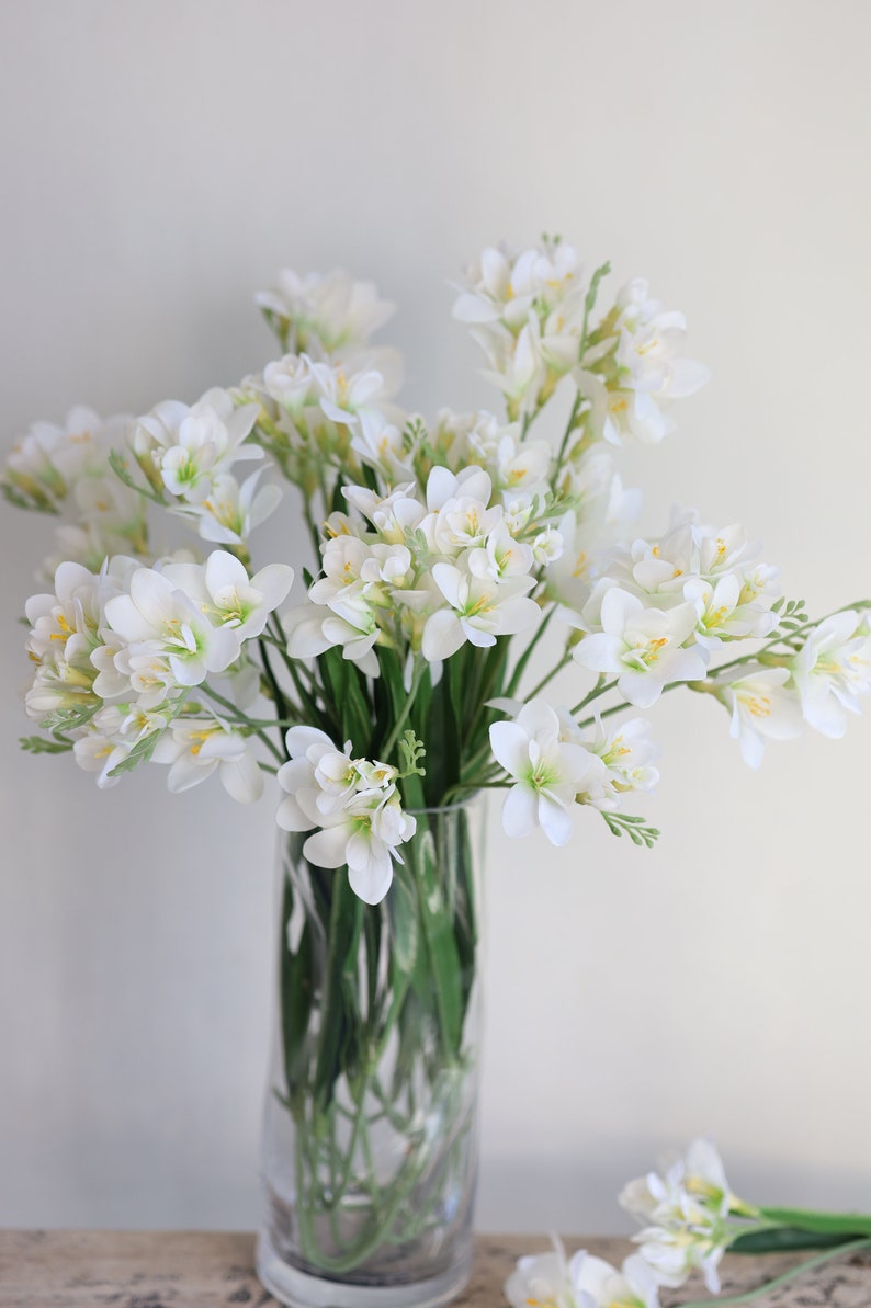 May include: A bouquet of white freesia flowers with yellow centers arranged in a clear glass vase. The flowers are in full bloom and have a delicate, sweet fragrance.