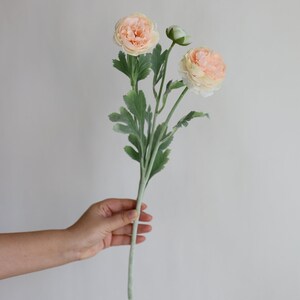 Puede incluir: Un ramo de tres flores artificiales de ran&uacute;nculo de color melocot&oacute;n con hojas y tallos verdes.