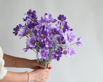 May include: A bouquet of purple cosmos flowers with yellow centers and green stems. The flowers are held in hands against a light gray background. The cosmos flowers have multiple petals and are in full bloom.