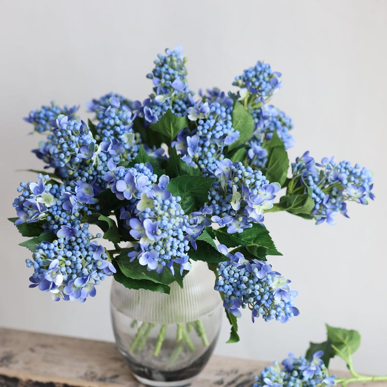 May include: A bouquet of blue artificial hydrangea flowers in a clear glass vase. The flowers are arranged in a cluster and have a realistic appearance.