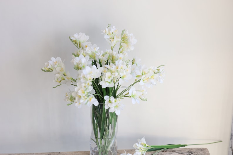 May include: A bouquet of white freesia flowers in a clear glass vase. The flowers are arranged in a loose, natural style.