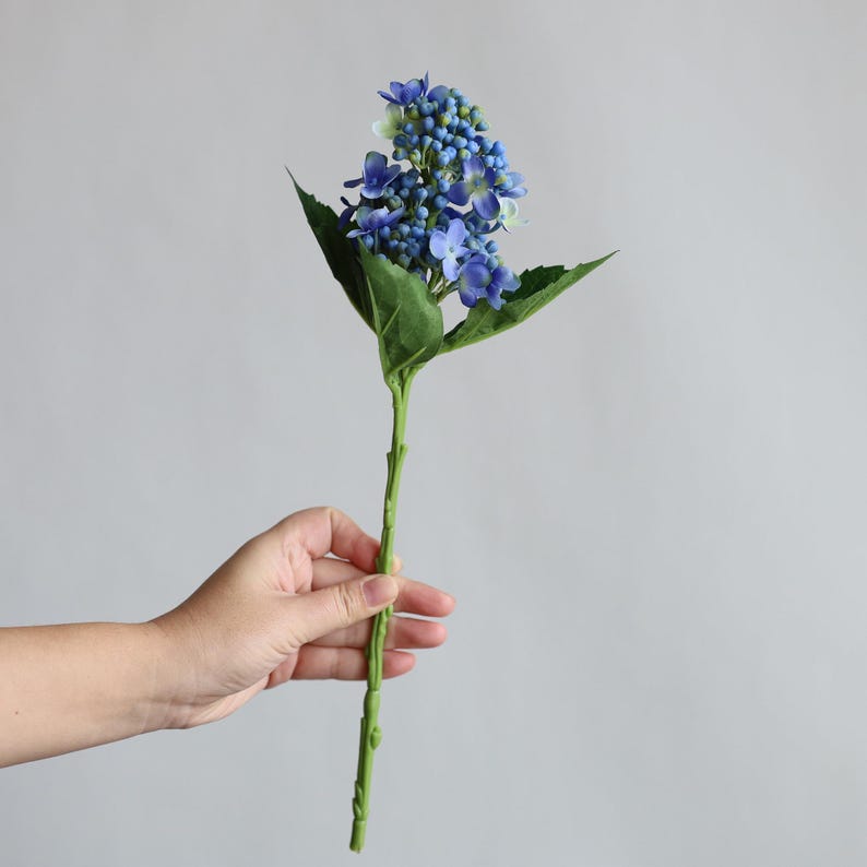May include: A single stem of artificial blue hydrangea flowers with green leaves.