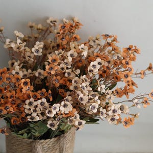 Puede incluir: Un ramo de flores artificiales con pequeñas flores delicadas en tonos crema y naranja. Las flores están dispuestas en una cesta tejida.