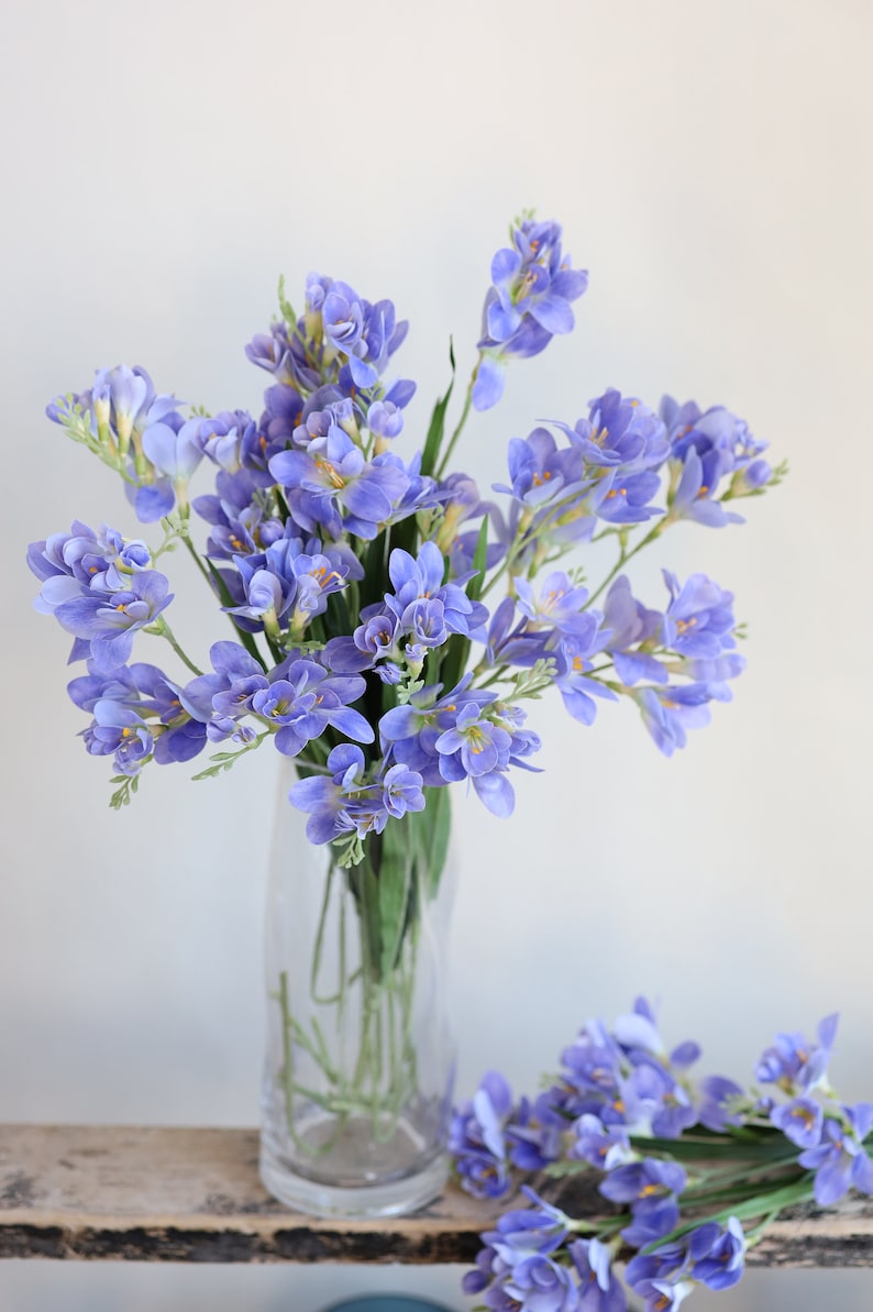 Puede incluir: Un ramo de flores de fresia moradas en un jarr&oacute;n de vidrio transparente. Las flores est&aacute;n dispuestas en un estilo suelto y natural.