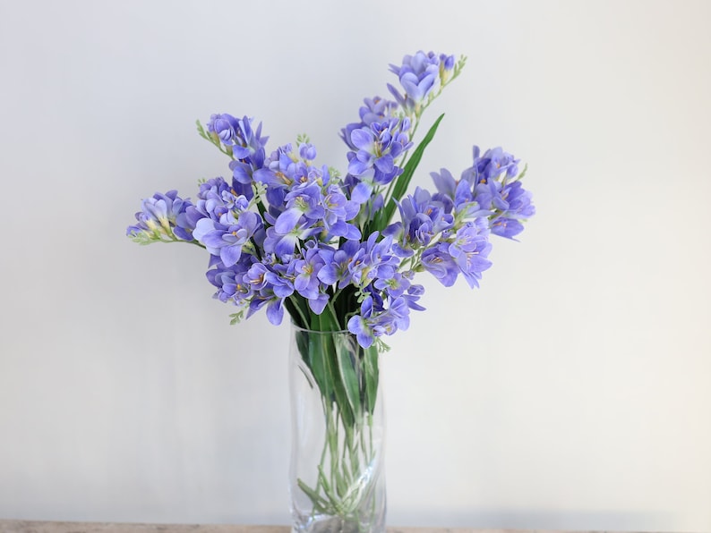 Puede incluir: Un ramo de flores de fresia moradas en un jarr&oacute;n de vidrio transparente. Las flores est&aacute;n dispuestas en un estilo suelto y natural.
