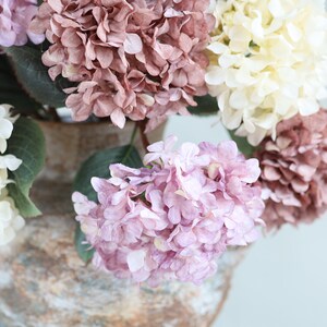 Puede incluir: Un ramo de flores de hortensia artificiales en un jarr&oacute;n r&uacute;stico. Las flores son en tonos de rosa, blanco y morado.
